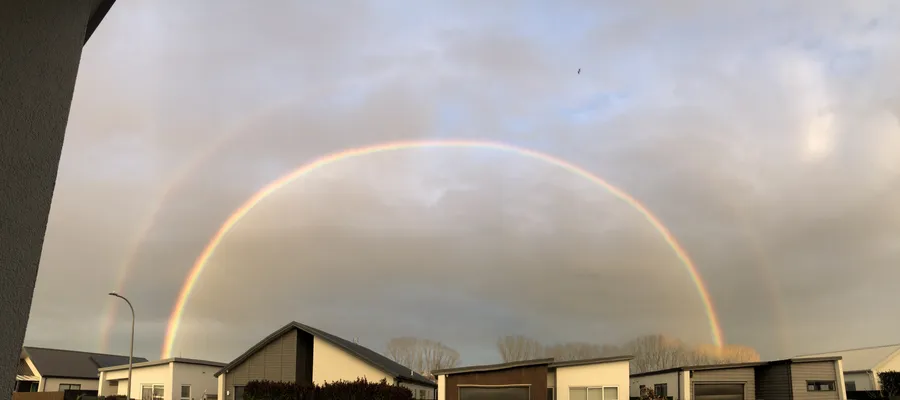 A full double rainbow arcing over a street of single storey houses under grey cloud, the fainter second bow sitting outside the bright one